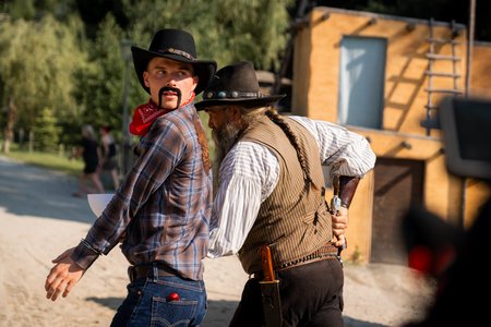 Zwei Männer in Cowboy-Kleidung sind in eine Szene verwickelt. Einer, mit falschem Schnurrbart, rotem Kopftuch und schwarzem Hut, blickt über seine Schulter zurück. Der andere, ein älterer Mann mit Bart, Weste und dunklem Hut, scheint ihm etwas zuzuflüstern, während er seine Schultern hält.