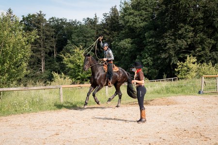 Eine Person auf einem Pferd wirft ein Lasso in die Luft, während eine Person am Boden zusieht und die Aktivität mit einem Smartphone aufzeichnet. Sie befinden sich in einem von Bäumen gesäumten Außenbereich mit einem Holzzaun und Kiesboden.