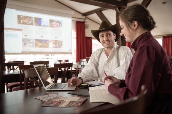 zwei Personen sitzen an einem Tisch mit Laptop und Flyer
