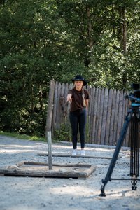 Person mit schwarzem Hut und dunkler Kleidung wirft eine Axt auf ein hölzernes Ziel im Freien, mit Bäumen und einem Holzzaun im Hintergrund. Im Vordergrund rechts ist die Kameraausrüstung zu sehen.