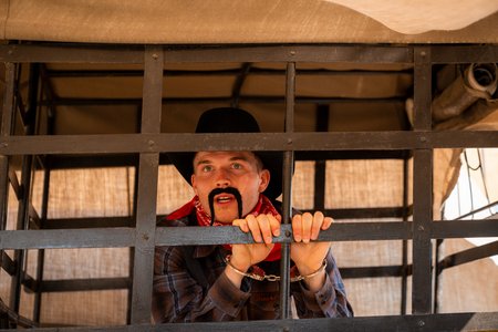 Eine als Cowboy verkleidete Person mit falschem schwarzen Schnurrbart, schwarzem Hut und rotem Bandana befindet sich in einer hölzernen Gefängniszelle, umklammert die Gitterstäbe mit beiden Händen und blickt hinaus. Im Hintergrund ist ein mit Plane bedeckter Wagen zu sehen.