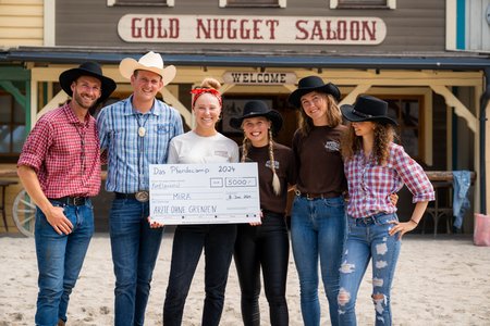 Eine Gruppe von sechs Personen in Westernkleidung steht vor einem Gebäude mit der Aufschrift „Gold Nugget Saloon“ in Pullman City. Sie halten einen großen Scheck über 5000 € für „Mina“ in den Händen. Die Gruppe wirkt glücklich und feierfreudig, einige Mitglieder tragen Cowboyhüte.