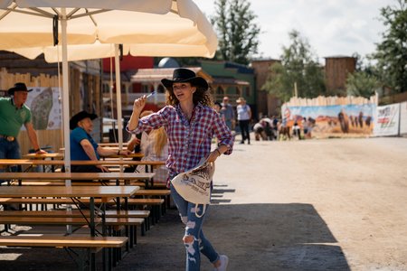 Eine Person in kariertem Hemd, Blue Jeans und Cowboyhut läuft fröhlich durch einen Außenbereich mit Holzbänken und Sonnenschirmen. Die Person trägt eine Tasche und zeigt nach oben. Im Hintergrund sind weitere Personen zu sehen, die sitzen und gehen.