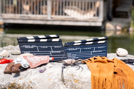 Zwei schwarze Filmklappen auf einer Steinoberfläche, umgeben von verschiedenen Filmrequisiten, darunter ein braunes Fransengewand, ein brauner Hut und eine Spielzeugpistole. Im Hintergrund von Pullman City sind eine Westernstadt-Holzkonstruktion und ein Gewässer zu sehen. Die Szene spielt im Freien an einem sonnigen Tag.