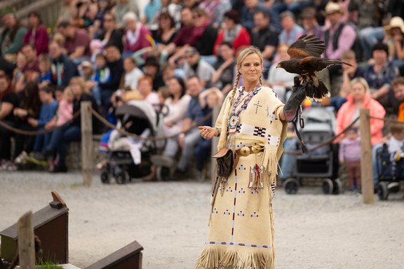 Eine Person in traditioneller Kleidung bietet ein spannendes Wild-West-Abenteuer, indem sie vor einem aufmerksamen Publikum im Freien eine Falknerei-Vorführung vorführt. Mit einem Greifvogel auf der ausgestreckten behandschuhten Hand wird die Menge von diesem außergewöhnlichen Erlebnis gefesselt.